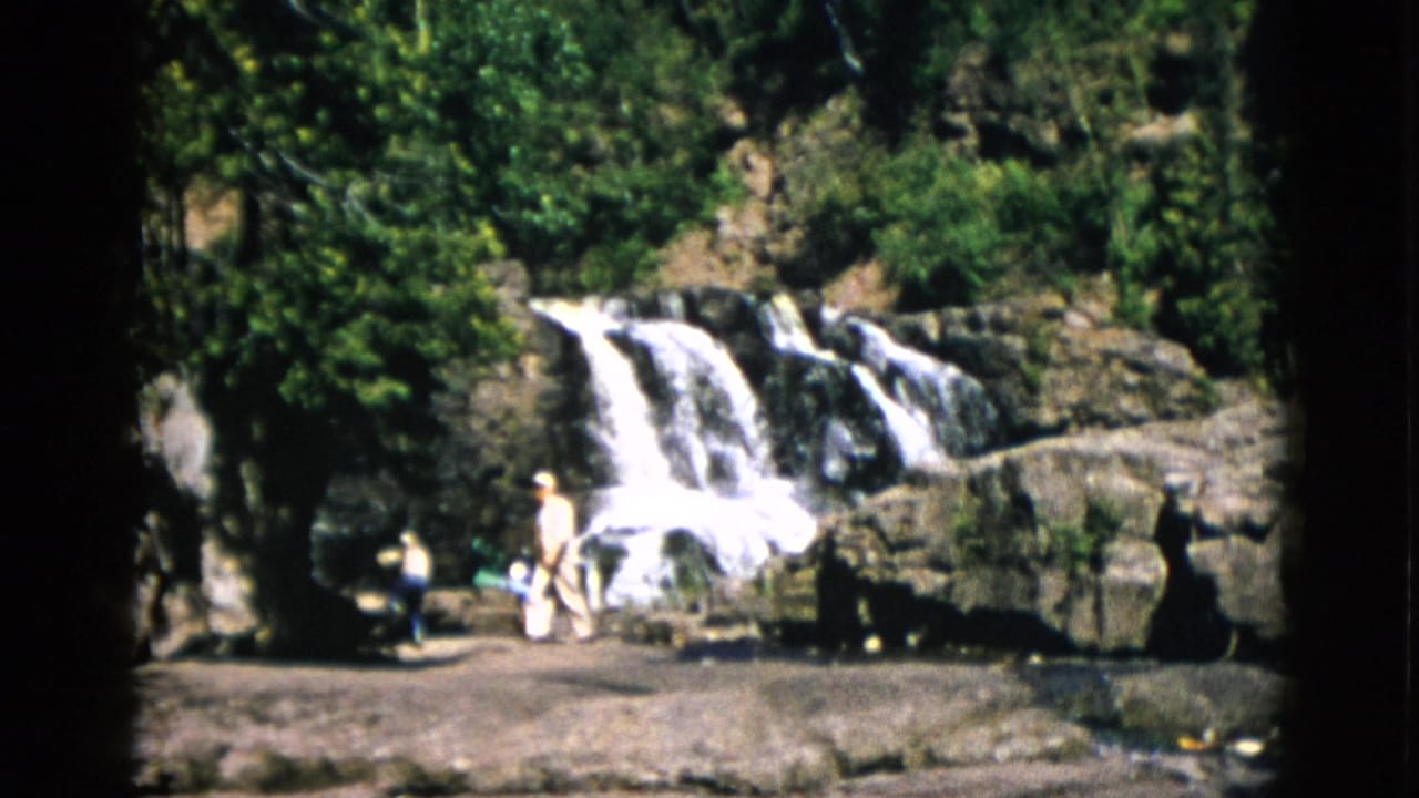 Cascading waterfall over dark rocky ledges with dense green trees; two or three figures at the base in light summer clothing.