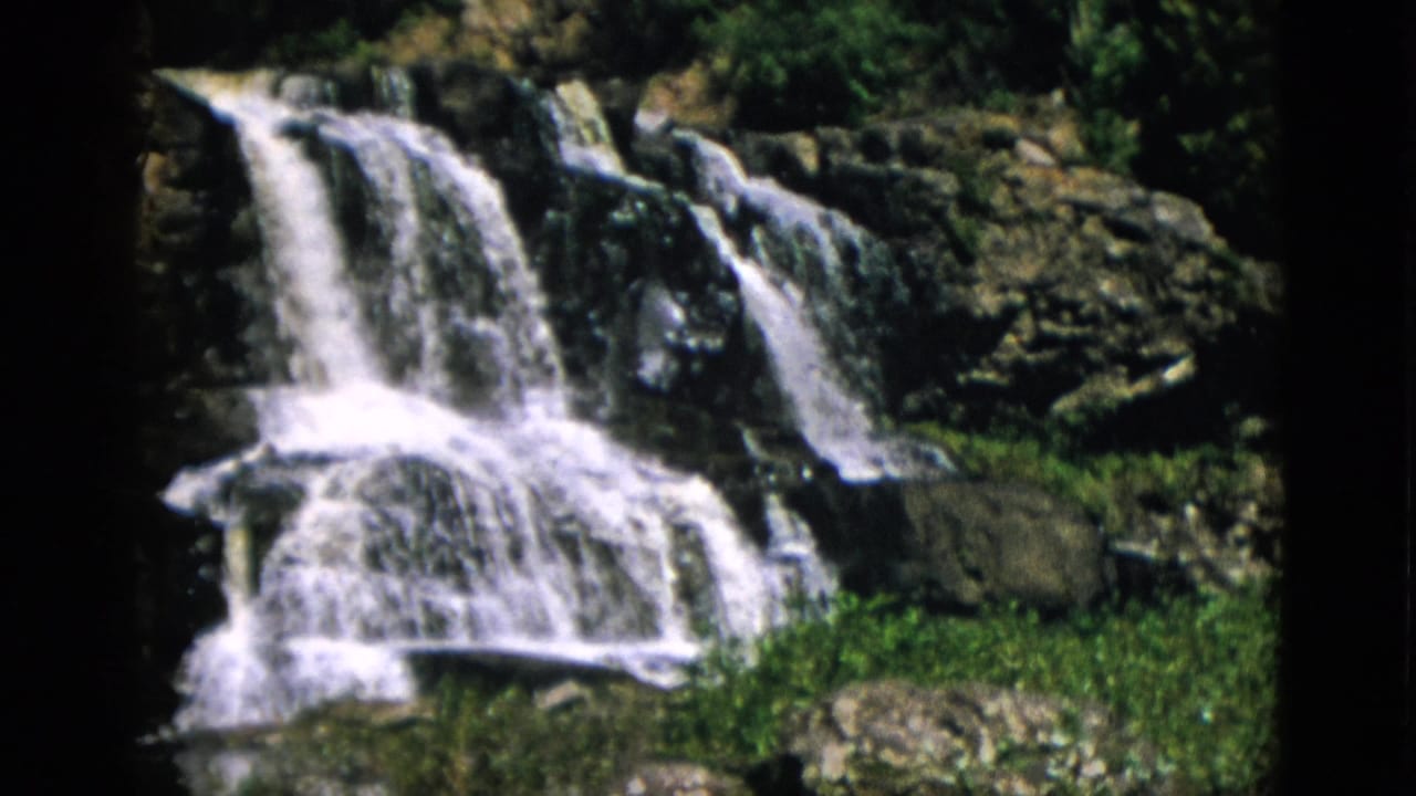 Waterfall flowing in multiple streams over dark rocky ledges, green ground cover at base, 8mm film.
