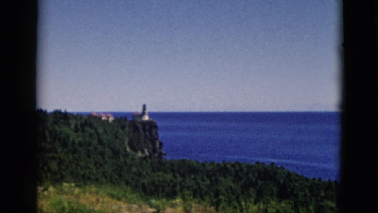 Opening 8mm frame from a 1952 Quebec home movie: a stone tower atop a rocky coastal cliff overlooking a large blue waterway, dense forest below, clear summer sky.