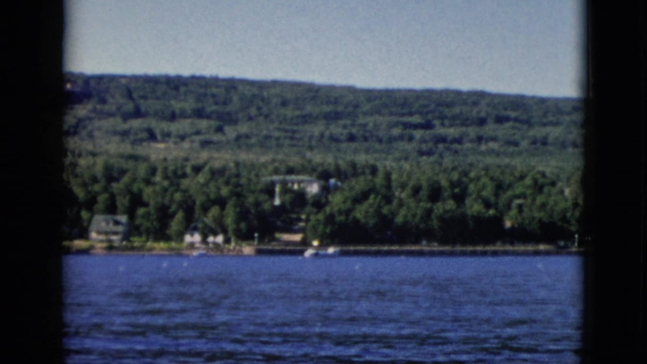1952 8mm home movie frame showing a wooded Canadian shoreline with period buildings partially obscured by dense forest and a broad waterway in the foreground.