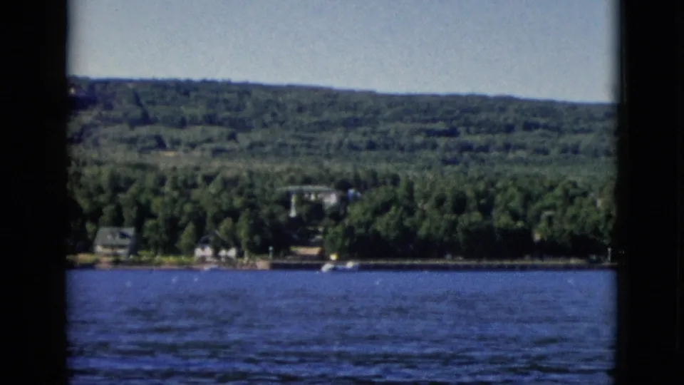 1952 8mm home movie frame showing a wooded Canadian shoreline with period buildings partially obscured by dense forest and a broad waterway in the foreground.