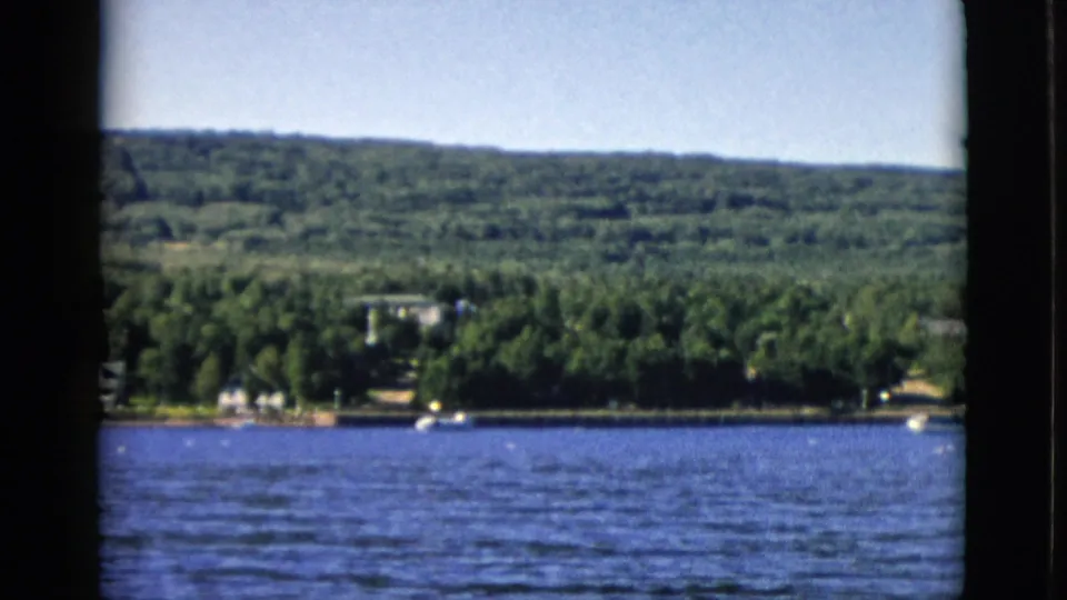 1952 8mm frame showing a stable wide shot of a forested Canadian shoreline, period buildings visible at the treeline, calm water surface under summer light.