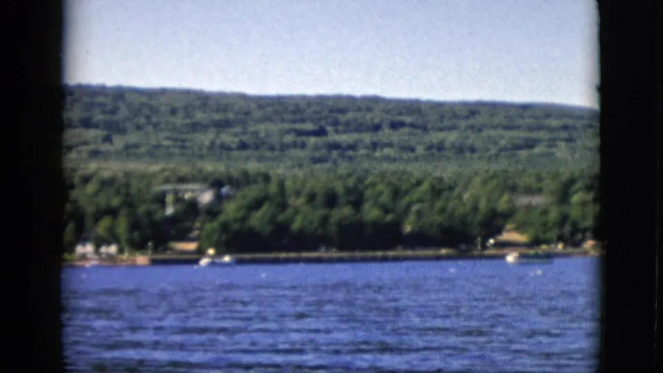 Closing 8mm frame of the 1952 Quebec shoreline sequence; wooded hillside, period buildings at water's edge, open waterway under a clear summer sky.