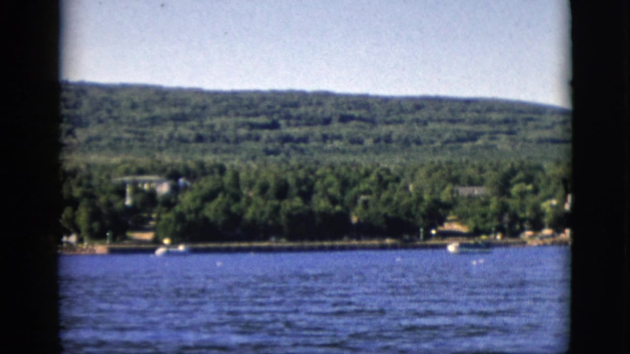 8mm color film, circa 1952, Quebec, Canada: blue lake foreground, dense deciduous forest and scattered residential structures on far shore, wooded hills behind.