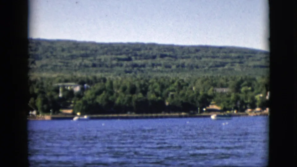 8mm color film, circa 1952, Quebec, Canada: blue lake foreground, dense deciduous forest and scattered residential structures on far shore, wooded hills behind.