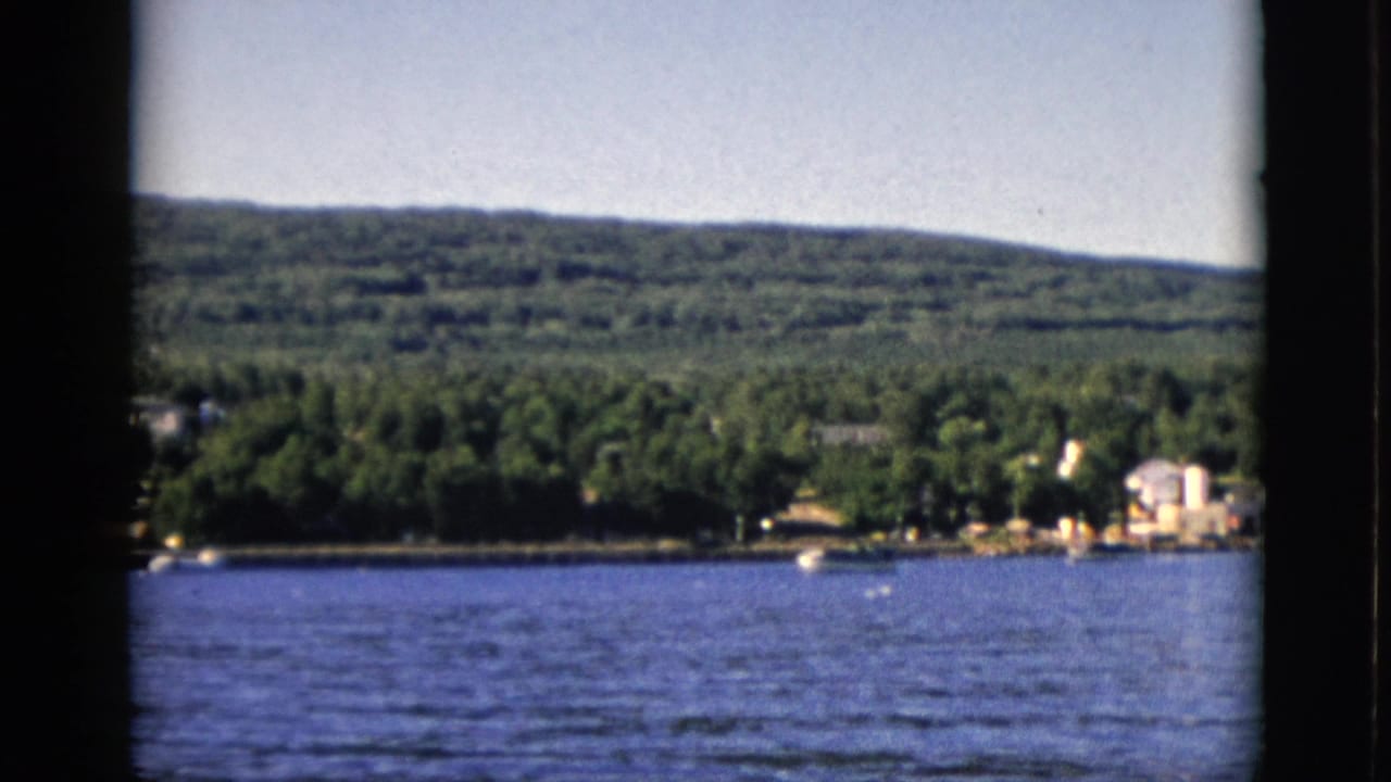 8mm color film, circa 1952, Quebec, Canada: lake foreground, far shore with trees and residential buildings on right, wooded hills under clear sky.