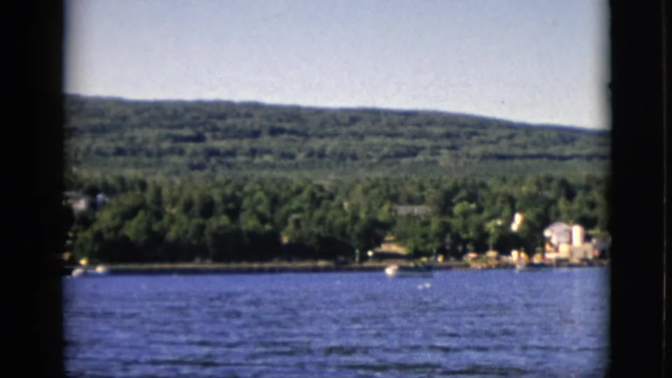 8mm color film, circa 1952, Quebec, Canada: lake foreground, far shore with trees and residential buildings on right, wooded hills under clear sky.