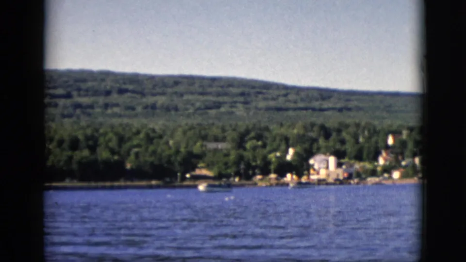 8mm color film, circa 1952, Quebec, Canada: rocky near-shore edge, blue lake, far shore with trees and buildings, wooded hills beyond.