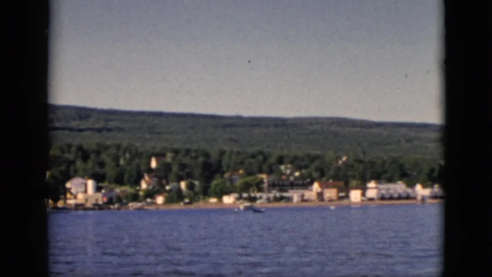 Reframed 8mm shot showing a broader sweep of the Quebec waterfront settlement, buildings spread along the shore with forest behind, circa 1952.