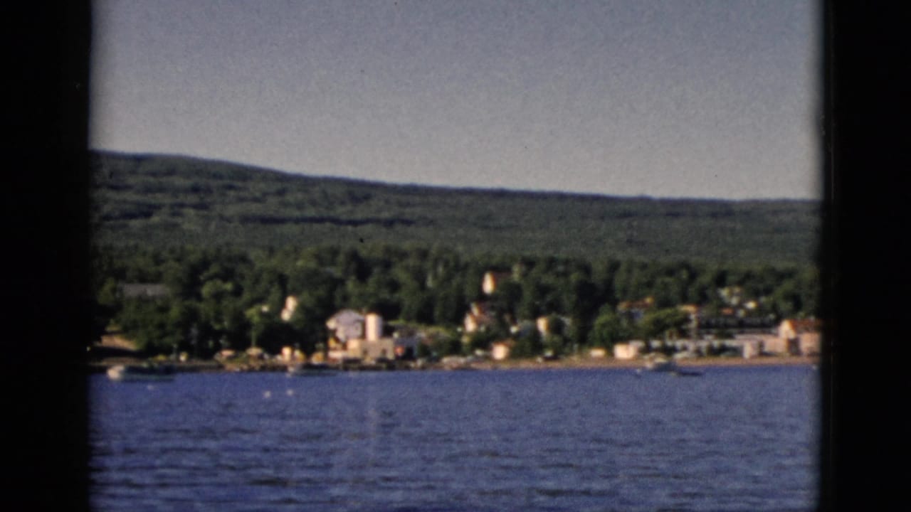 8mm color shot from water level looking toward a small Quebec lakeside settlement amid dense forest, with rolling hills beyond, circa 1952.