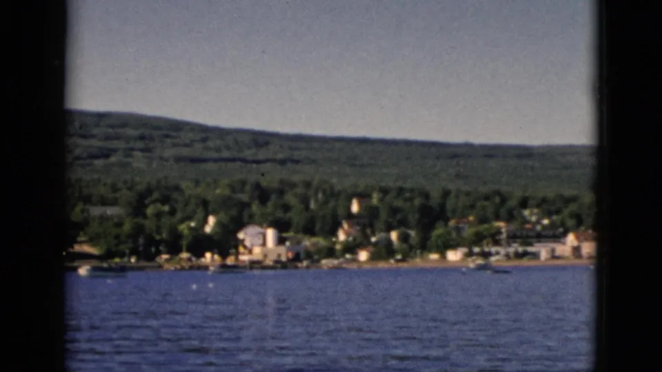 8mm color shot from water level looking toward a small Quebec lakeside settlement amid dense forest, with rolling hills beyond, circa 1952.