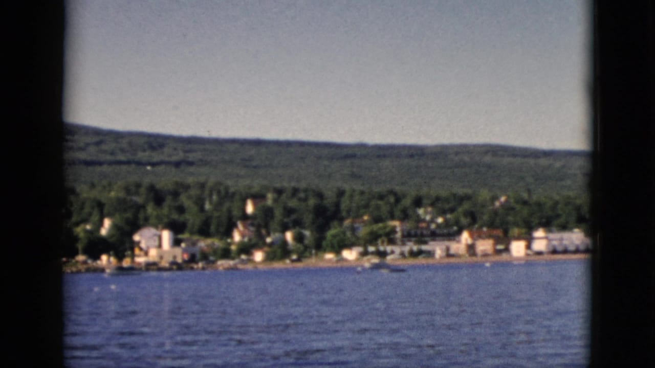 Second 8mm frame of a Quebec lakeside community from water level, showing modest buildings set against dense forest and gentle hills, circa 1952.
