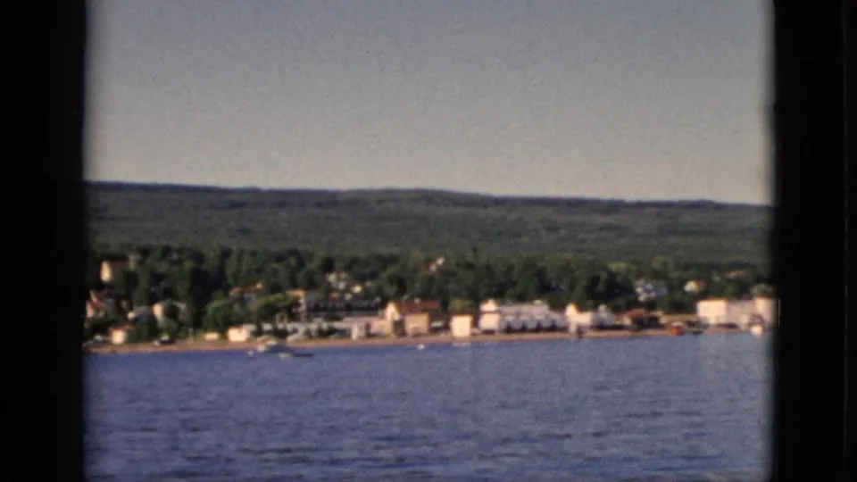 Closing 8mm frame of a small Quebec lakeside community from water, forested hills behind the settlement under a clear summer sky, circa 1952.