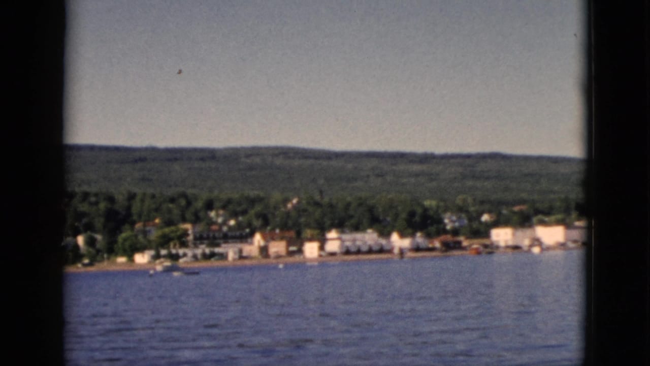 1952 8mm home movie frame showing a small Quebec lakeside settlement from across the water, with dense mixed forest and rolling hills behind.