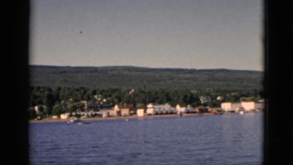 1952 8mm home movie frame showing a small Quebec lakeside settlement from across the water, with dense mixed forest and rolling hills behind.
