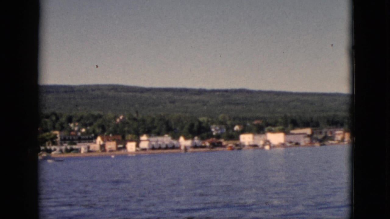 1952 8mm home movie of a Quebec lakeside town seen from water, with forested hills and rows of small white buildings along shore.