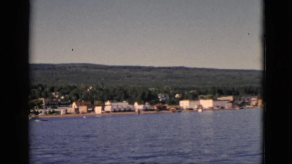 1952 8mm home movie of a Quebec lakeside town seen from water, with forested hills and rows of small white buildings along shore.