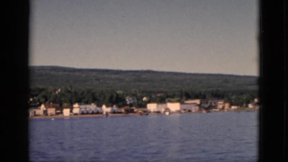 1952 Quebec home movie frame showing lakeside town buildings more prominently, with mixed forest rising behind the settlement.