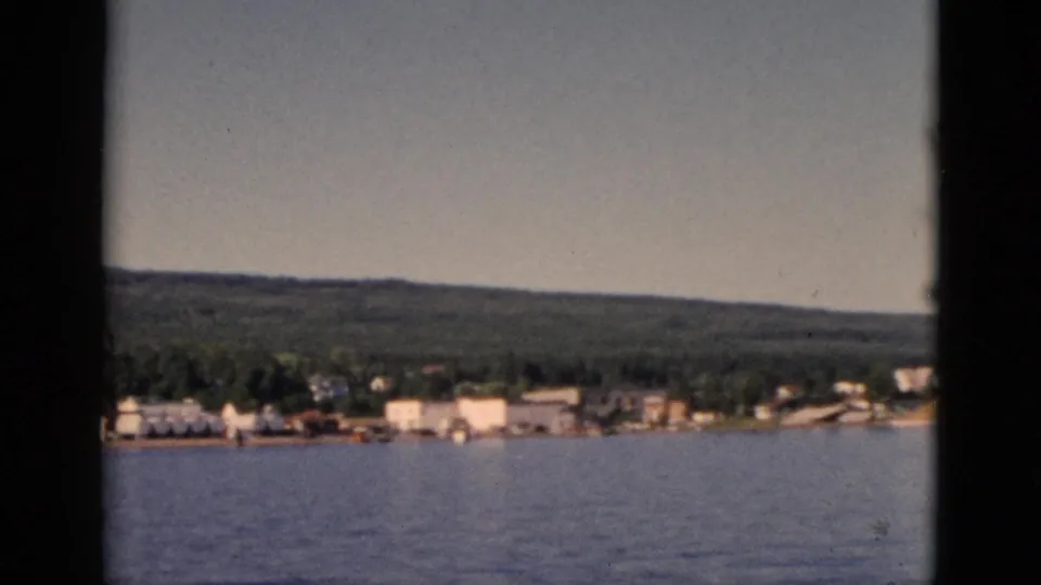 1952 8mm home movie frame of a small Quebec lakeside town with a possible dock structure at the shore and dense forested hills behind.