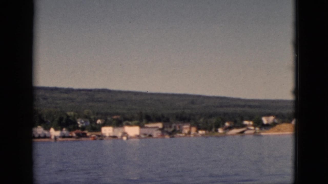 View from a moving boat of a small lakeside settlement with white buildings and a forested hill, circa 1952, Quebec.