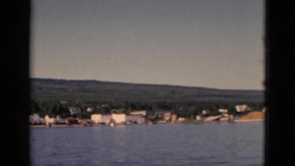 8mm footage circa 1952 — a small Quebec lakeside community seen from water level; white buildings line the shore beneath a dense forested hillside.