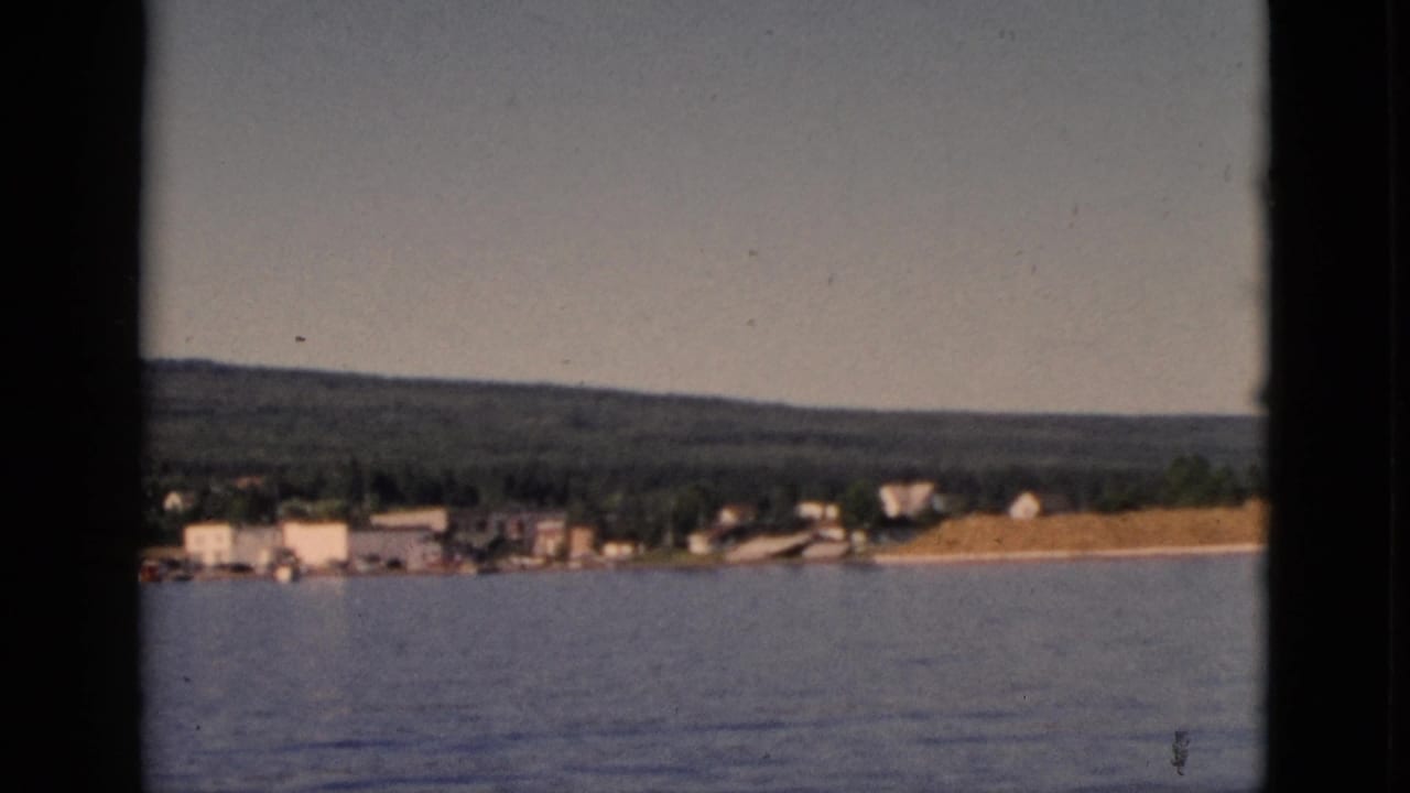 Boat-POV of Quebec shoreline with clustered white buildings, sandy beach strip, and forested hill, circa 1952.