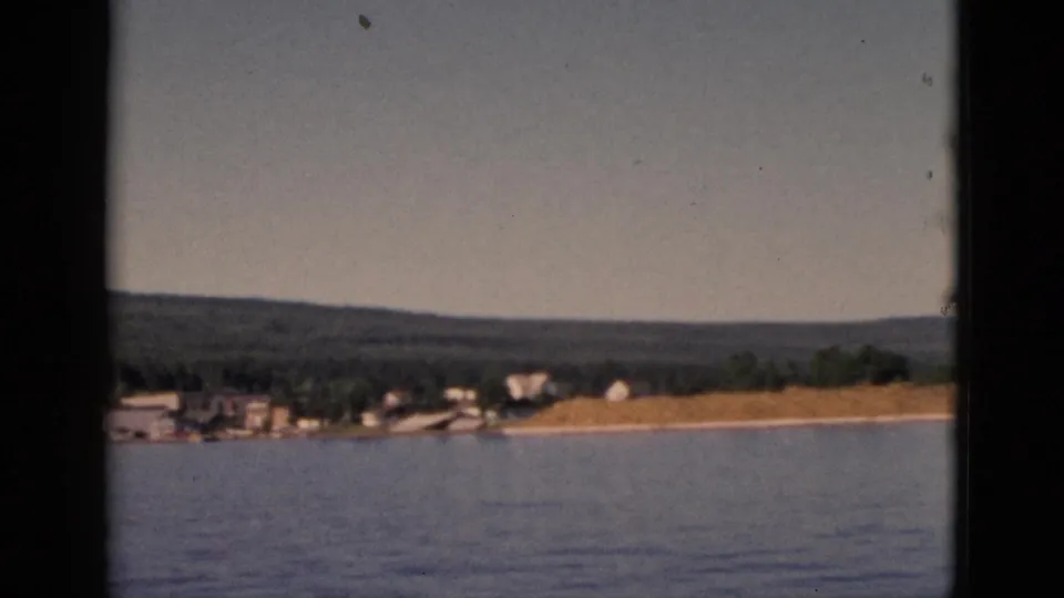 8mm frame circa 1952, Quebec — camera has panned further along the shore, revealing a sandy embankment and sparse buildings under a broad forested hillside.