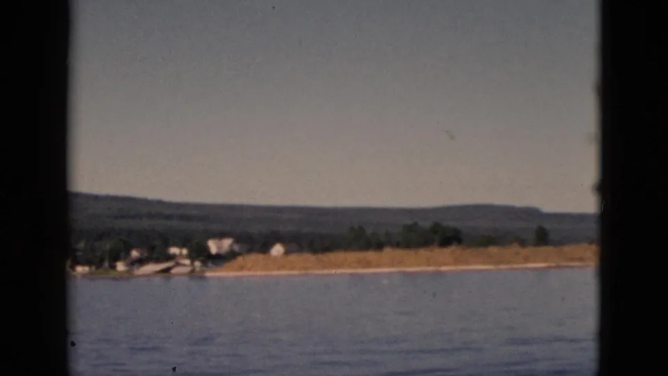 8mm footage near clip end, circa 1952 Quebec — shoreline buildings and sandy bank remain visible as the boat continues past the small settlement.