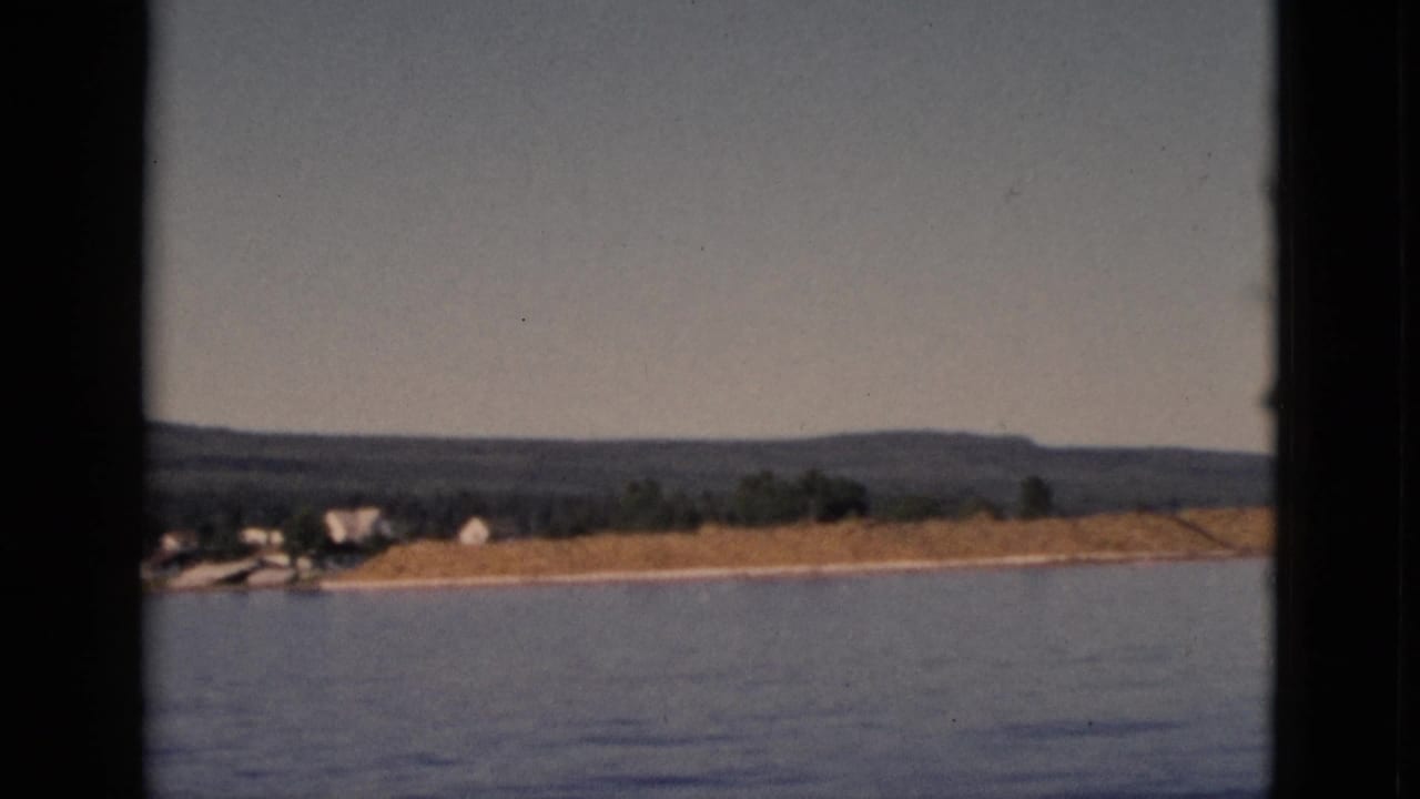 Calm lake water in foreground with distant sandy shoreline, white house, treeline, and rolling hills under pale sky.