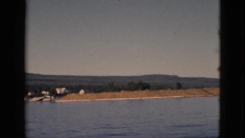 View from a moving boat across calm water toward a low sandy shore with scattered white houses and forested hills, Quebec, Canada, circa 1952.
