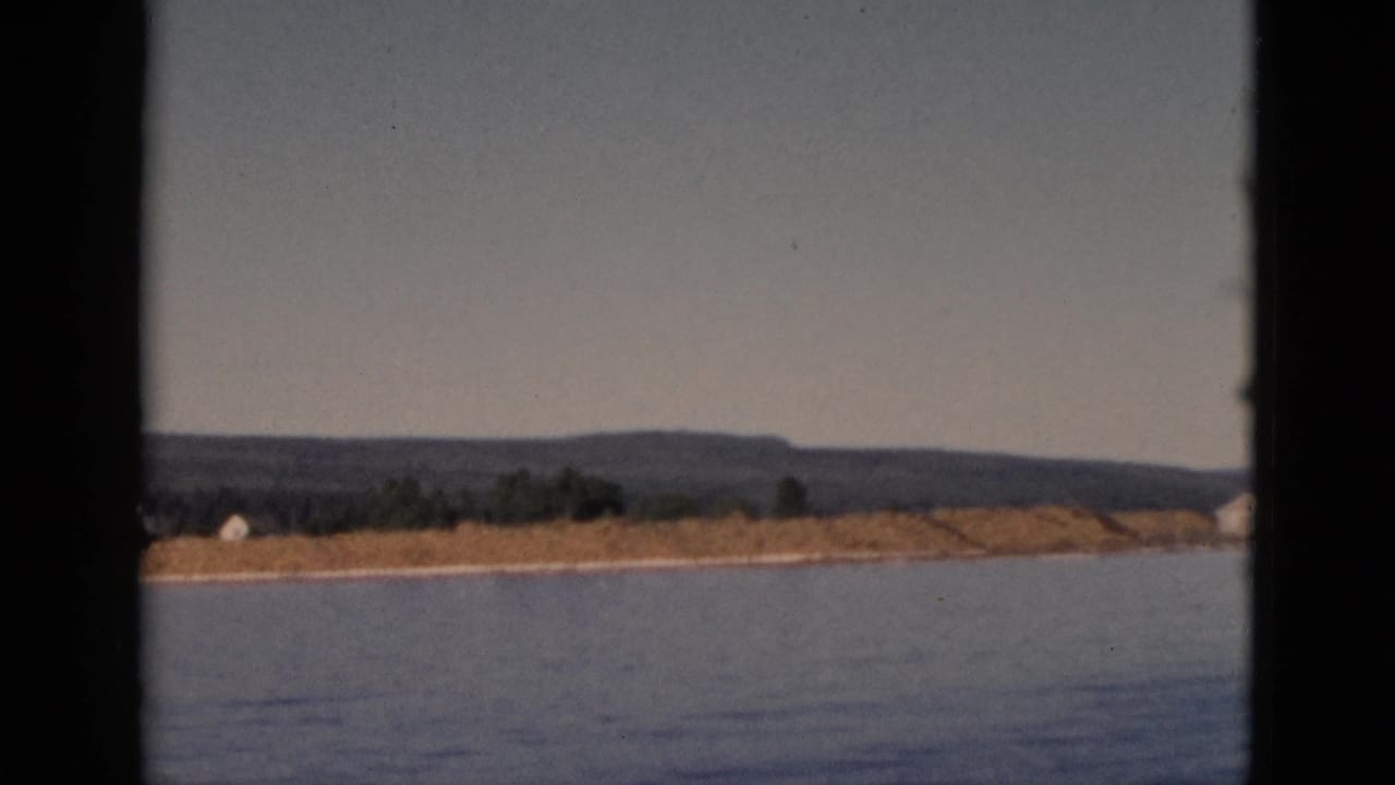 Boat-POV shot across still water toward a low sandy shore with a white building, tree cover, and hills, Quebec, Canada, circa 1952.