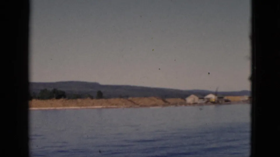 Moving boat view of calm water, low shore with small scattered structures, trees, and hills, Quebec, Canada, circa 1952.