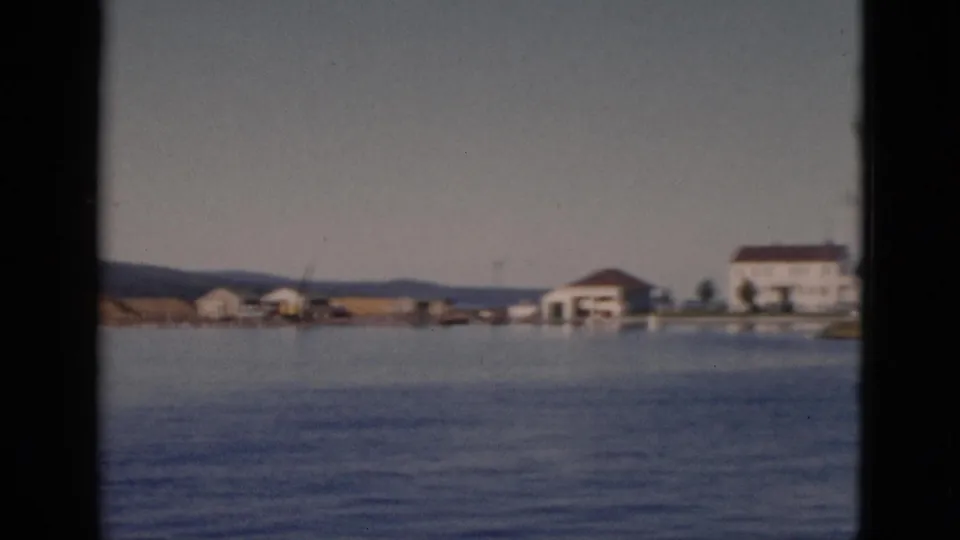 Boat POV reveals a cluster of white buildings with a prominent larger structure with a dark roof on a low Quebec shoreline, circa 1952.