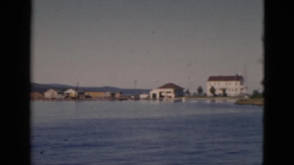 8mm color footage, c.1952, Quebec, Canada — calm water in foreground, white multi-story building with red-brown roof and adjacent waterfront structures on a distant shore.