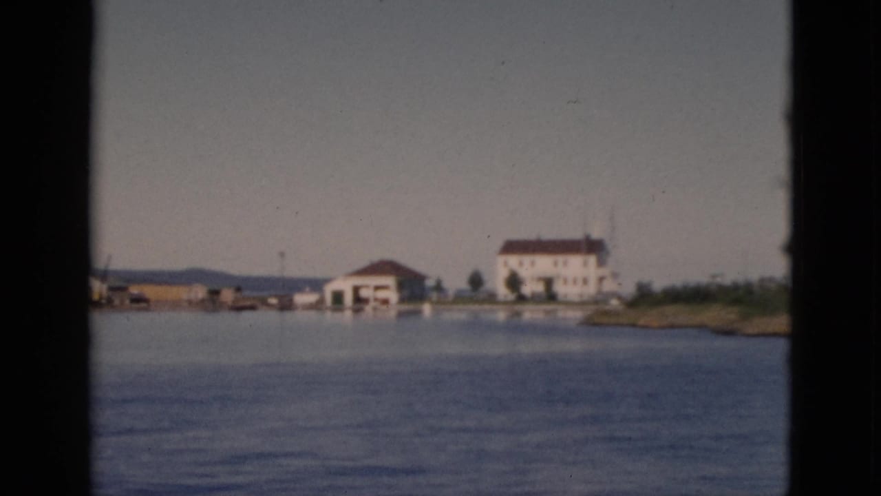 Continued boat-side view of the Quebec shoreline; white building with red-brown roof prominent at center.