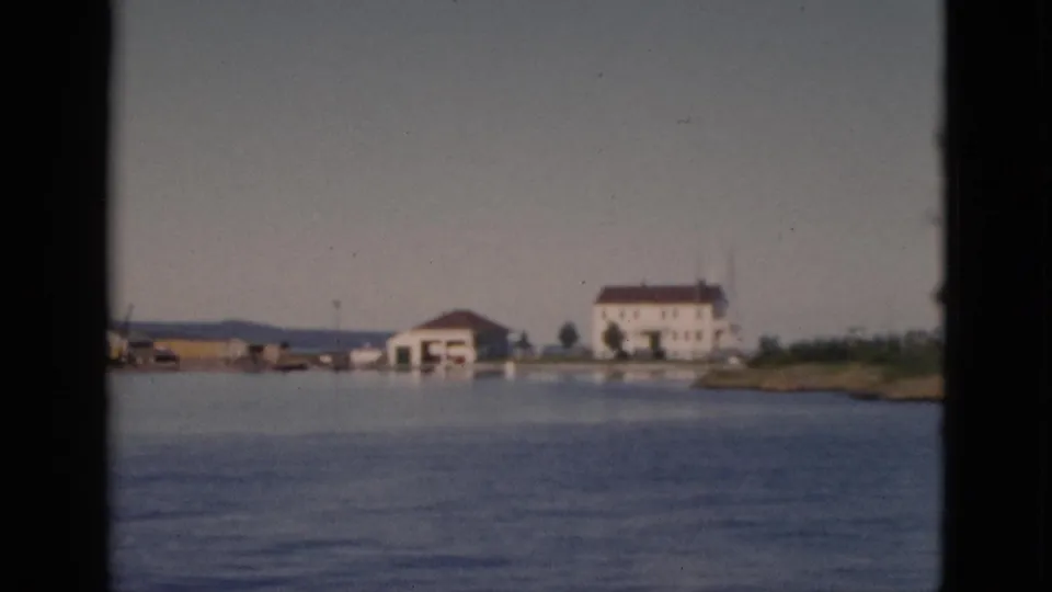 8mm color footage, c.1952, Quebec — boat drifts past a waterfront cluster of white buildings, the largest with a red-brown roof, on calm water.