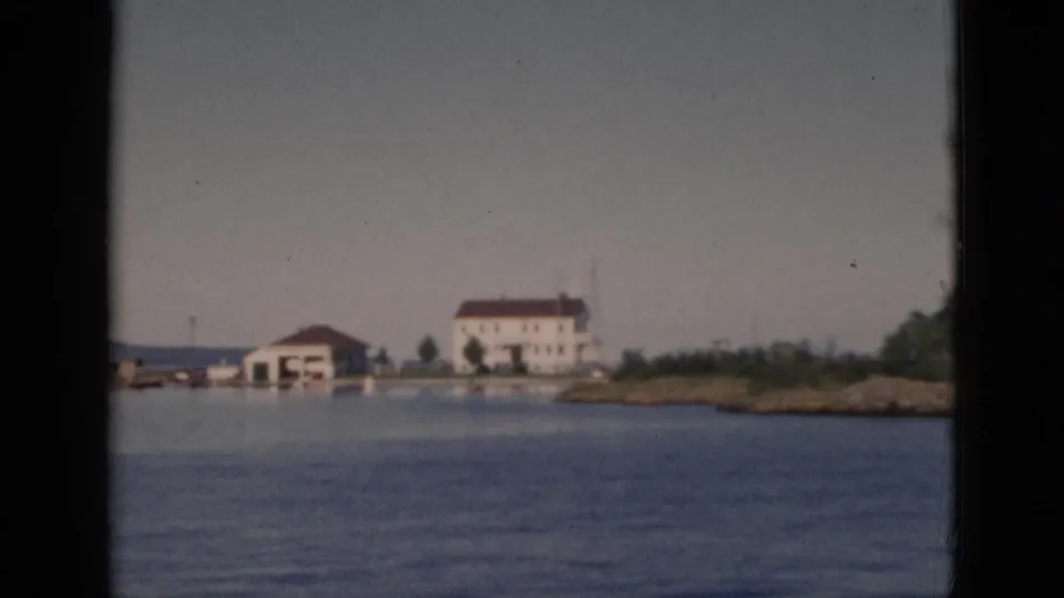 8mm color footage, c.1952, Quebec — rocky low-lying shoreline enters foreground as boat moves closer; white multi-story building with red-brown roof remains central.