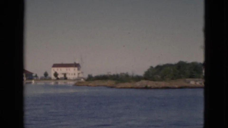 8mm color footage, c.1952, Quebec — densely wooded island or promontory enters frame at right as boat continues past; white building with red-brown roof at center-left.