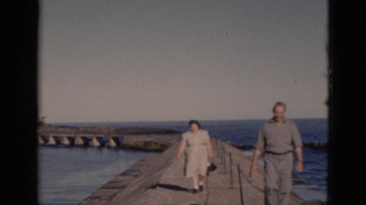 Couple in mid-century casual dress walking forward along a narrow concrete pier with rope railing, calm open water on both sides, Quebec home movie circa 1952.