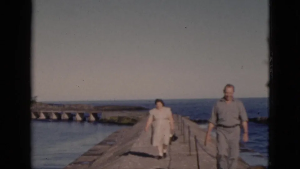 Couple in mid-century casual dress walking forward along a narrow concrete pier with rope railing, calm open water on both sides, Quebec home movie circa 1952.