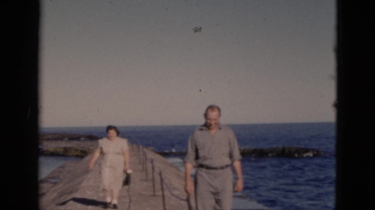 Couple advancing toward camera on a concrete pier; man in belted trousers and short-sleeve shirt, woman in light dress, Quebec home movie circa 1952.