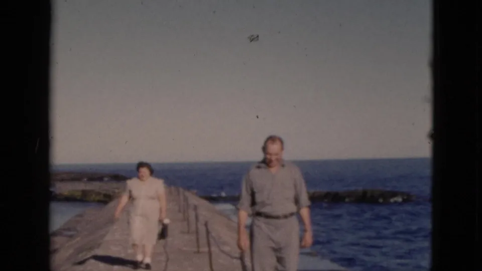 Couple advancing toward camera on a concrete pier; man in belted trousers and short-sleeve shirt, woman in light dress, Quebec home movie circa 1952.