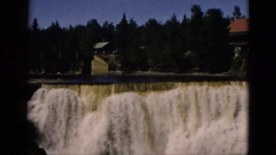 Broad waterfall over a wide rocky ledge with a red-roofed building and dense evergreen treeline above; consistent with a Quebec tourist site, home movie circa 1952.