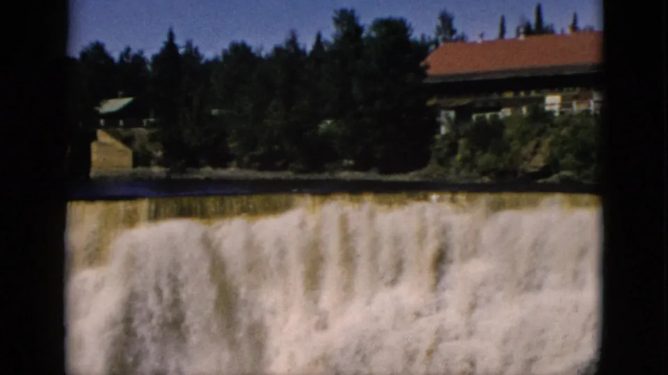 Continued wide shot of broad waterfall and red-roofed building with dense evergreen treeline, Quebec home movie circa 1952.