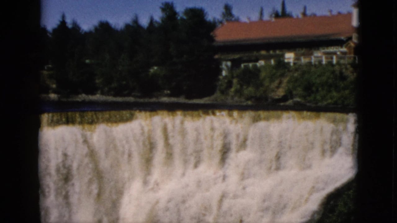8mm home movie frame, c. 1952, Quebec, Canada: wide cascading waterfall with a brick building with red roof on the cliff edge above the falls and dense conifers.