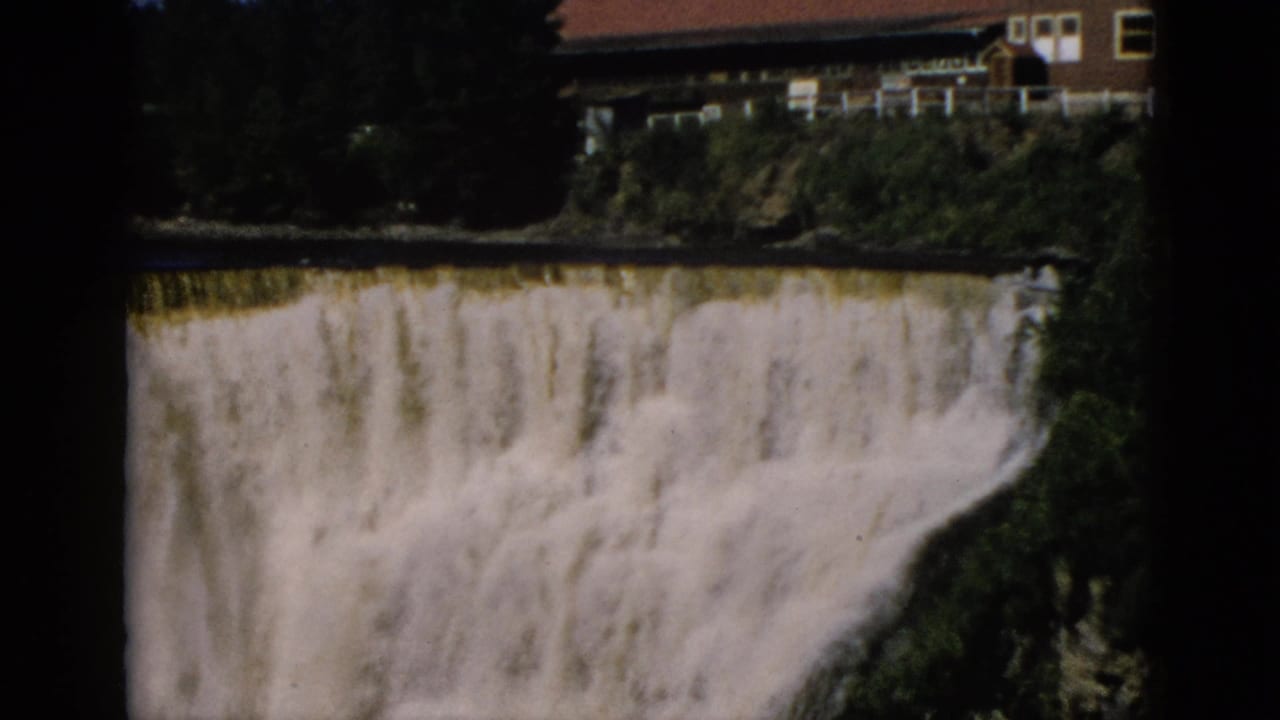 Cascading waterfall with a multi-story building with red roof on the cliff top; possible film notation artifact visible in upper right corner.