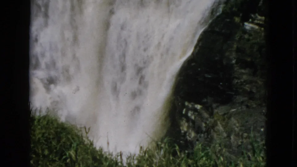Large waterfall beside dark rock face; green grass and vegetation appear at lower right, 8mm home movie circa 1952, Quebec, Canada.