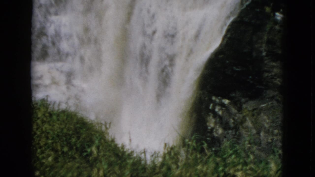 Cascading waterfall over dark rocks with green grassy mound in foreground, 8mm home movie frame