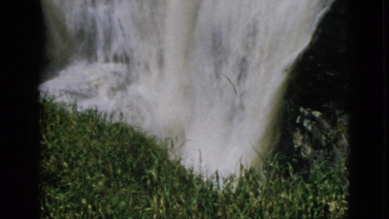 Waterfall beside green mossy embankment with dark rock, 8mm home movie frame