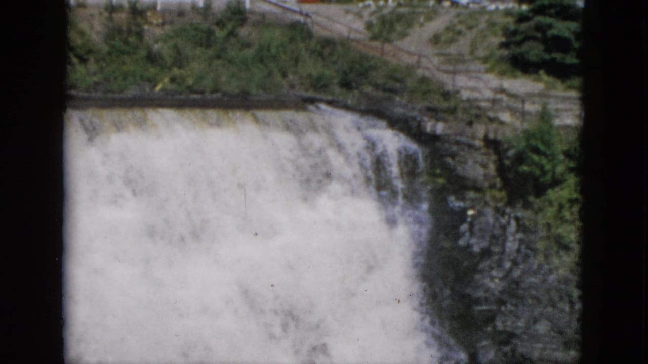 1950s 8mm color film: wide view of a powerful waterfall in Quebec, Canada, with green hillside, dark layered cliff, and roadside infrastructure at the crest.